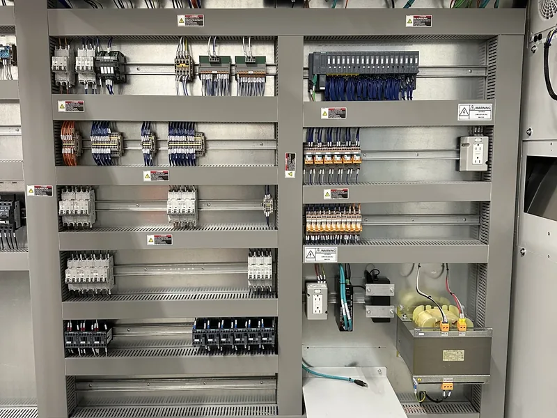 Particolare dell'armadio: moduli del PLC, protezioni e trasformatore, con le etichette di avvertenza